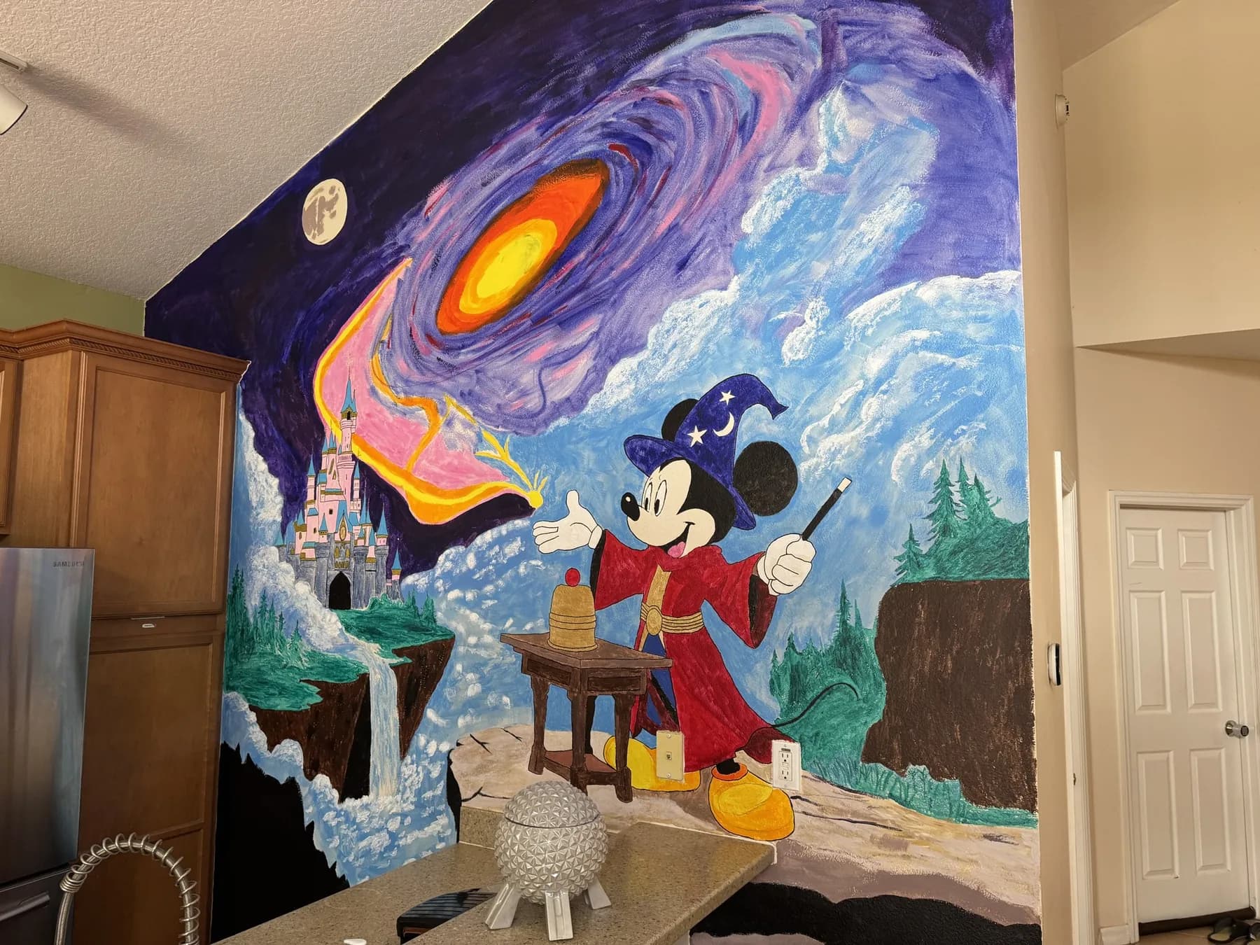 The finished mural on a tall kitchen wall: a galaxy and clouds above Mickey Mouse as the Sorcerer's Apprentice, doing a cups trick at a small table, with a castle and a waterfall on the left